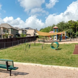a fenced in dog park with a bench and a playground in the background